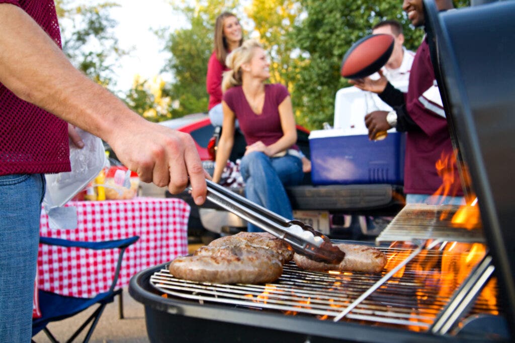 Man tossing burgers on a charcoal grill. People in background tossing football having a tailgate party.