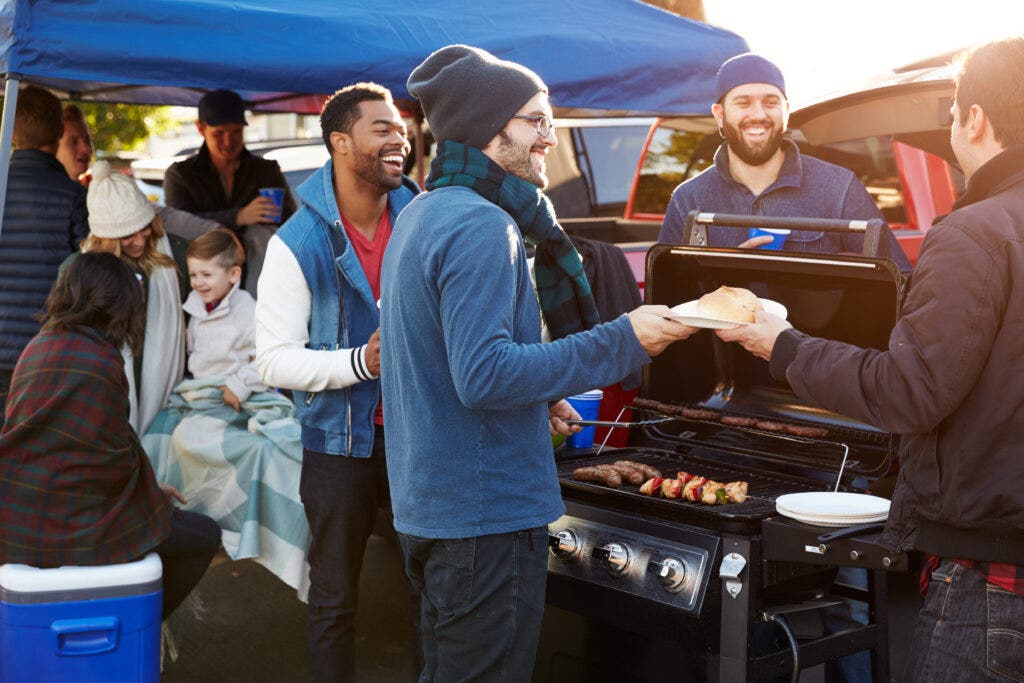 Group of fans tailgating underneath a blue canopy pop-up party tent. Gathered around black grill. Wearing Blue winter hats having a tailgate party. 